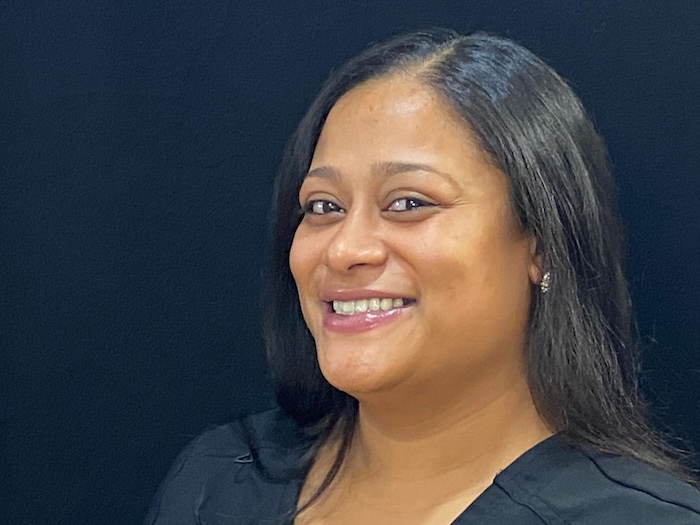 The image is a portrait of a woman smiling at the camera, with a friendly and welcoming expression. She has dark hair, appears to be wearing a black top or dress, and is positioned against a neutral background that does not distract from her.