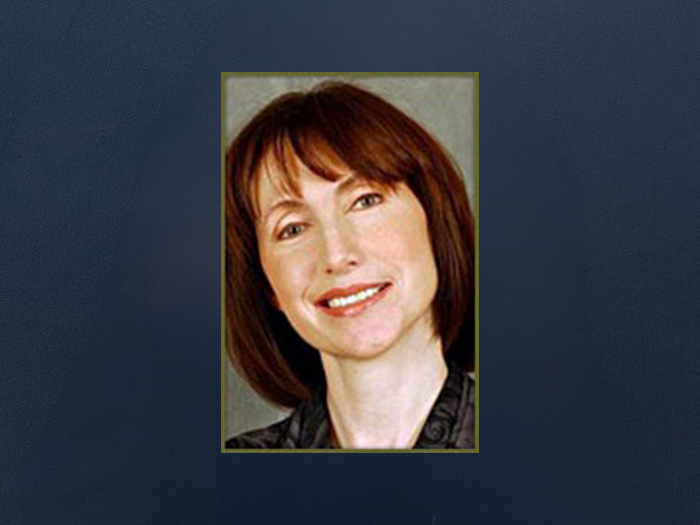 The image is a portrait of a woman with short hair, smiling slightly and looking directly at the camera. She has fair skin and appears to be middle-aged or older. The woman is wearing a dark top and her expression is friendly and approachable.