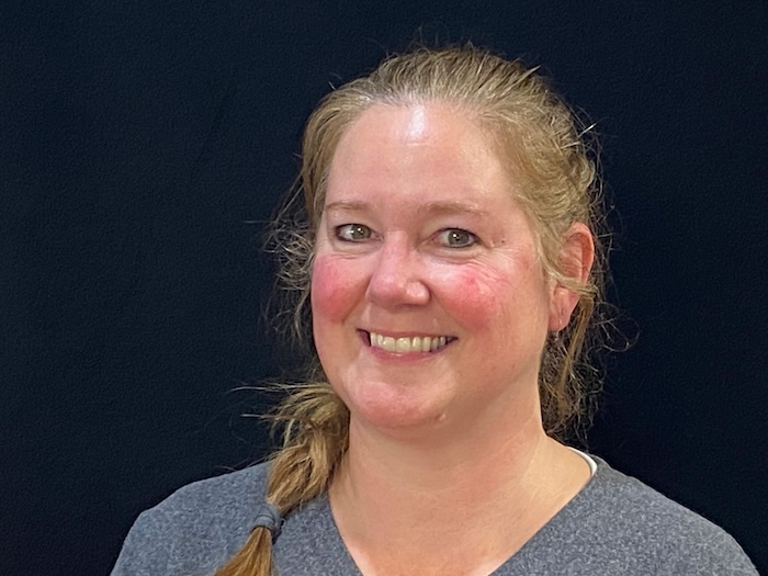 A smiling woman with a beard, wearing a gray top and posing against a dark background.