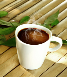 A white coffee cup with steam rising from the top, placed on a wooden surface alongside some fresh green leaves.