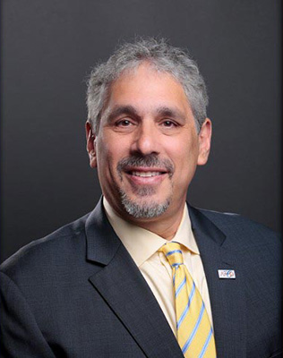 The image is a photograph of a smiling man with graying hair, wearing a suit and tie, standing against a dark background.