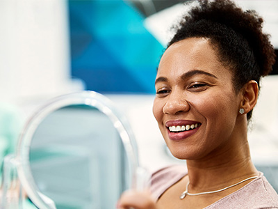 A woman with a bright smile is seated in front of a mirror, smiling broadly at the camera.