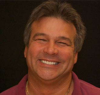 The image shows a man smiling at the camera, with short hair and wearing a dark t-shirt.