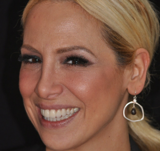 The image shows a woman with short blonde hair, wearing large hoop earrings and a smile. She has a light complexion and is looking directly at the camera.
