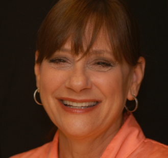 The image is a portrait of a smiling woman with short brown hair, wearing a pink top and large hoop earrings.