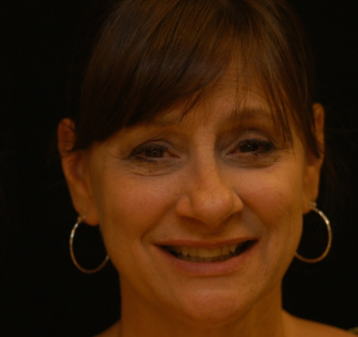 The image shows a woman with short hair, wearing a hoop earring and a smile. She has fair skin and appears to be indoors with a dark background.