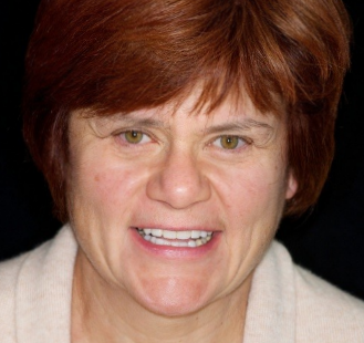 The image shows a woman with short hair and glasses, wearing a red top. She has a neutral expression and is looking directly at the camera.