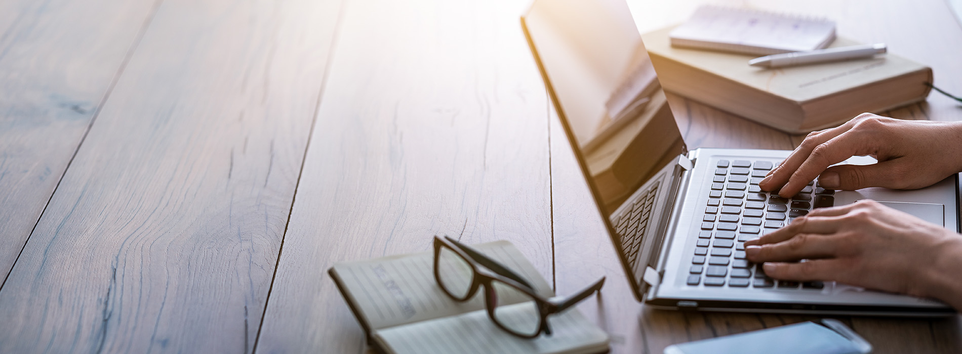 A person typing on a laptop with their hands, surrounded by books and papers, against a blurred background.