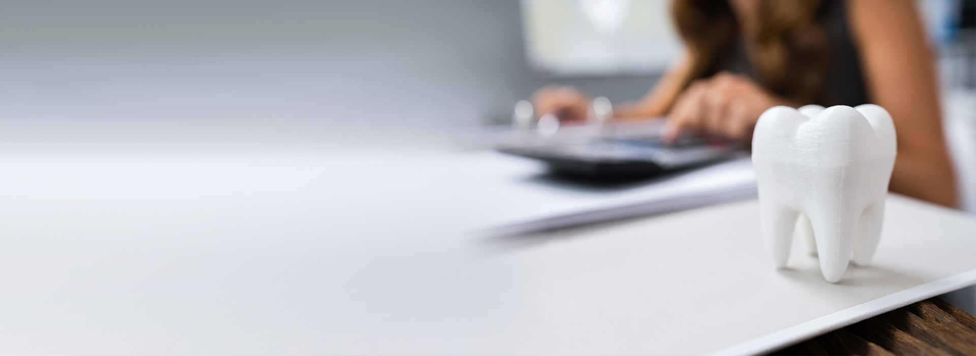 The image shows a person working at a desk with a computer monitor, keyboard, and mouse. In the foreground, there is a small white figurine of a dental implant.