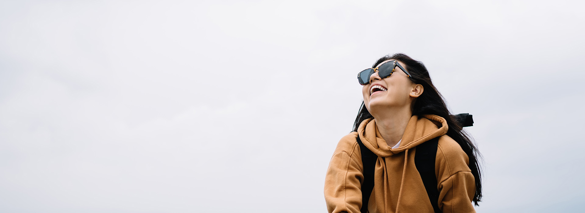 A person, possibly a woman, is smiling and looking upwards while standing outdoors with sunglasses on. She is wearing a backpack and a hoodie, which suggests casual or travel attire. The background is blurred but appears to be an overcast sky.