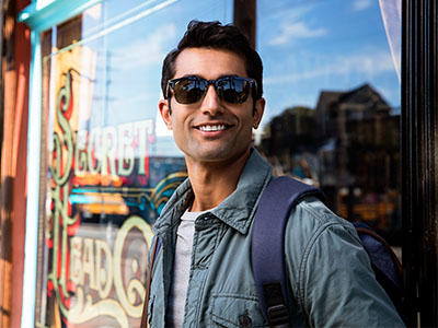 A young man in sunglasses and a backpack, standing outdoors in front of a storefront.