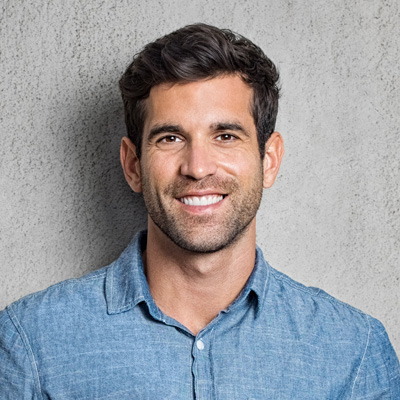 A smiling man with short hair, wearing a blue denim shirt and standing against a neutral gray background.