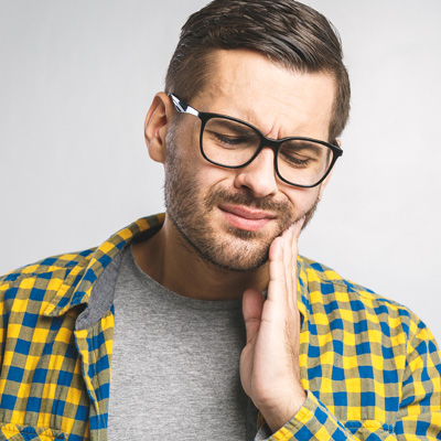 A man with a beard, wearing glasses and a yellow plaid shirt, is holding his hand to his mouth, possibly in pain or discomfort.
