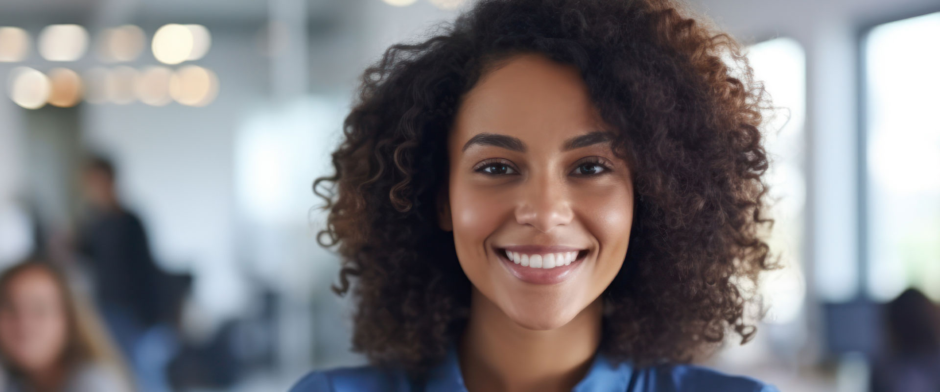 The image shows a smiling woman with curly hair, wearing a blue top and standing in an office environment.