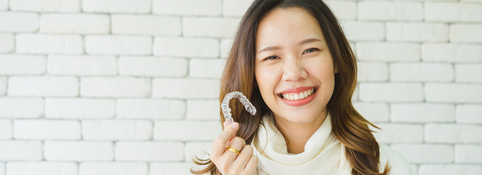 The image is a photograph of a smiling woman wearing a ring and holding a small object, set against a blurred background with a brick wall and white tiles.