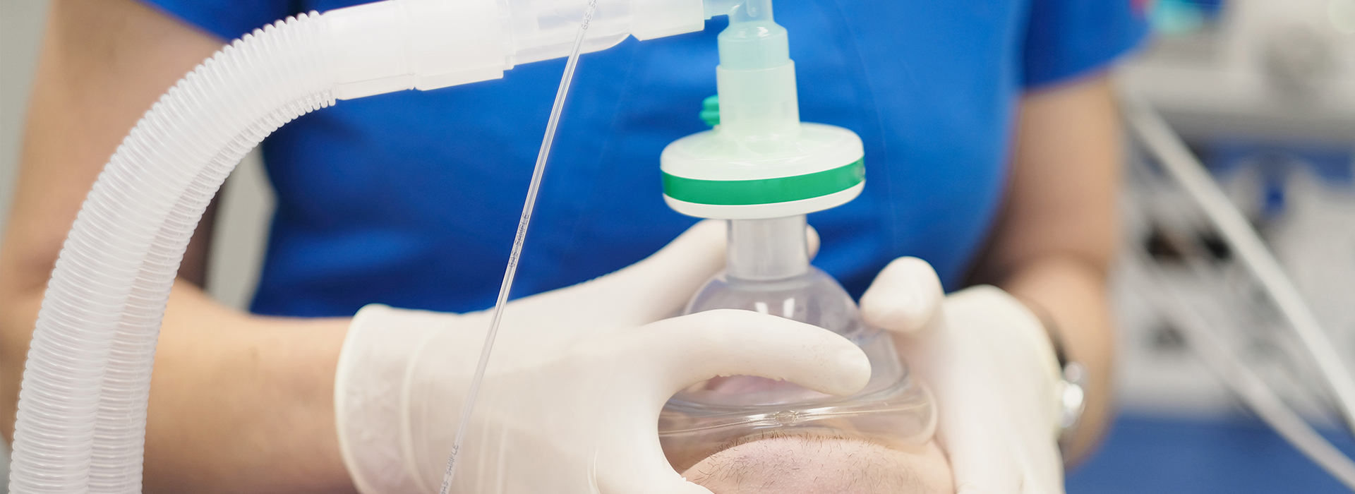 A person in a blue shirt and white gloves is holding a clear plastic bottle with a green cap, placed on a machine with tubes connected to it.