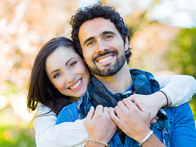 A man and a woman embrace, smiling at the camera, with the man wearing a bandana around his neck.