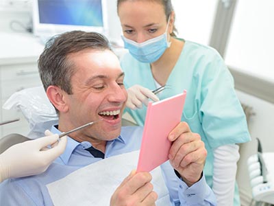 A man is sitting in a dental chair, holding a pink card with a surprised expression while a dentist and hygienist look on.