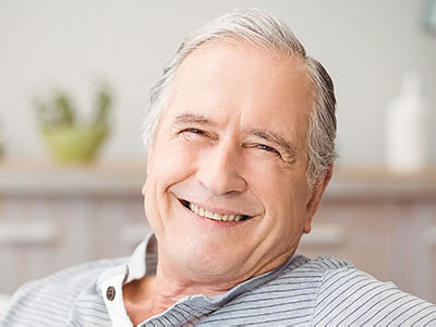 The image shows a smiling older man with gray hair, wearing glasses and a blue shirt, sitting comfortably in a chair indoors.