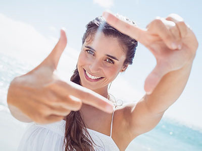 The image shows a woman taking a selfie with her hand held up to the camera, smiling at it. She is wearing a white top and has long hair. The background suggests a beach setting with clear skies.